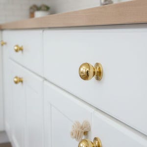 May include: Close-up of white kitchen cabinets with gold-colored spherical knobs. The cabinets have a classic, paneled design. A light-colored wooden countertop is visible above the cabinets. A small, decorative, beige-colored tassel is attached to one of the knobs.