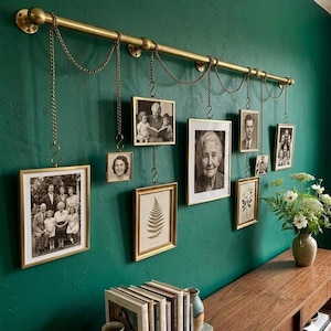 May include: A brass-colored wall-mounted photo display with hanging frames. The frames hold black and white photos and are suspended by chains. The wall is painted a deep teal green, and a wooden console table is below.