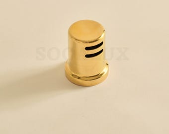 May include: A gold-colored, cylindrical bottle stopper with two horizontal slits near the top. The stopper has a rounded top and a flared base. The background is a solid, light beige color.