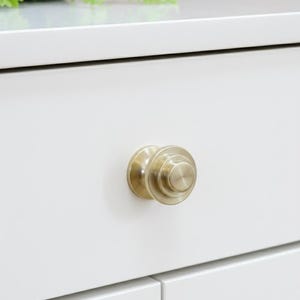 Peut inclure: Gros plan sur une armoire blanche avec un bouton de tiroir rond de couleur or. Le bouton de tiroir a un design en couches et est centré sur l'armoire. L'armoire fait partie d'un meuble plus grand.