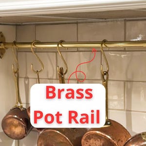May include: A brass pot rail with several S-hooks holding copper pots. The rail is mounted on a tiled wall. The text "Brass Pot Rail" is displayed in a white box with red lettering.