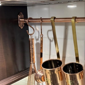May include: A copper-colored kitchen utensil rack with hanging tools. The rack is mounted on a white tiled wall and features a copper rod with various kitchen tools, including a whisk and measuring cups. The rack is approximately 24 inches long.