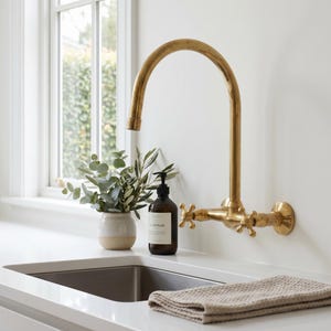 Peut inclure: Un robinet de cuisine doré avec des poignées en croix est monté sur un mur blanc. Un distributeur de savon brun foncé et un petit vase avec de la verdure sont posés sur le comptoir. Une serviette beige pliée repose près de l'évier en acier inoxydable.