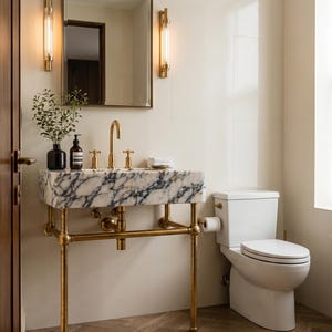 Peut inclure: Salle de bain luxueuse avec un lavabo en marbre et des robinets en laiton, soutenu par un support en laiton. Des toilettes blanches sont à proximité. Les murs sont neutres et le sol a un motif imitation bois. Deux appliques murales encadrent un grand miroir.