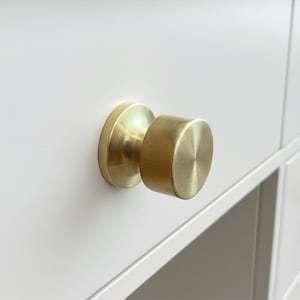 Peut inclure: Gros plan d'un bouton de meuble de couleur dorée. Le bouton a une base circulaire et une poignée cylindrique. La quincaillerie est montée sur une porte d'armoire blanche. Le bouton a une finition en métal brossé.