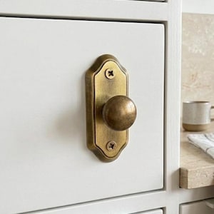 Peut inclure: Gros plan sur un bouton de meuble en laiton avec une poignée ronde et une plaque arrière rectangulaire. Le bouton est monté sur une porte d'armoire blanche. Le matériel a une esthétique vintage.