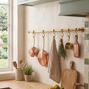 Peut inclure: Une barre de cuisine en laiton fixée au mur avec des crochets en S supportant deux casseroles en cuivre, un torchon en lin beige, un bouquet d'herbes séchées, une tasse à mesurer en laiton et une râpe en bois. Une bouilloire en cuivre est posée.