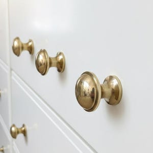 Peut inclure: Gros plan sur une commode blanche avec plusieurs poignées de tiroir dorées. Les poignées ont une forme arrondie en forme de champignon et sont espacées uniformément sur les tiroirs. La commode semble être en bois.