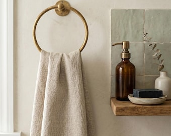 Puede incluir: Una toalla de lino beige cuelga de un toallero dorado en una pared clara. Una estantería de madera sostiene un dispensador de jabón marrón, una pastilla de jabón negro y un pequeño jarrón blanco. Decoración de baño minimalista.