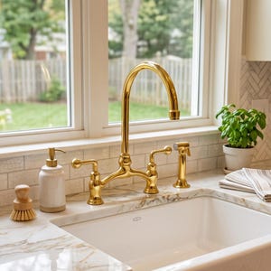 May include: A polished gold kitchen tap with a high arch spout, two lever handles, and a matching side sprayer. The tap is installed on a white marble countertop next to a white ceramic soap dispenser and a wooden dish scrubbing brush.