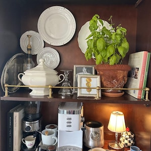 May include: A dark wood hutch shelf featuring a white tureen, a green basil plant in a terracotta pot, a white espresso machine, a small lamp, books, and various kitchenware including white plates, a silver tray, and a framed olive illustration.