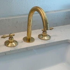 May include: A gold-coloured bathroom tap set with a curved spout and two separate handles. The tap is mounted on a white countertop above a white sink. The drain stopper is also gold-coloured.