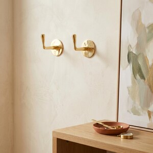 May include: Two brass wall hooks with a hammered finish are mounted on a light-colored wall. The hooks have a simple, elegant design, perfect for hanging coats, bags, or other items. A small, round, terracotta-colored bowl and a gold-colored watch are on a wooden surface.