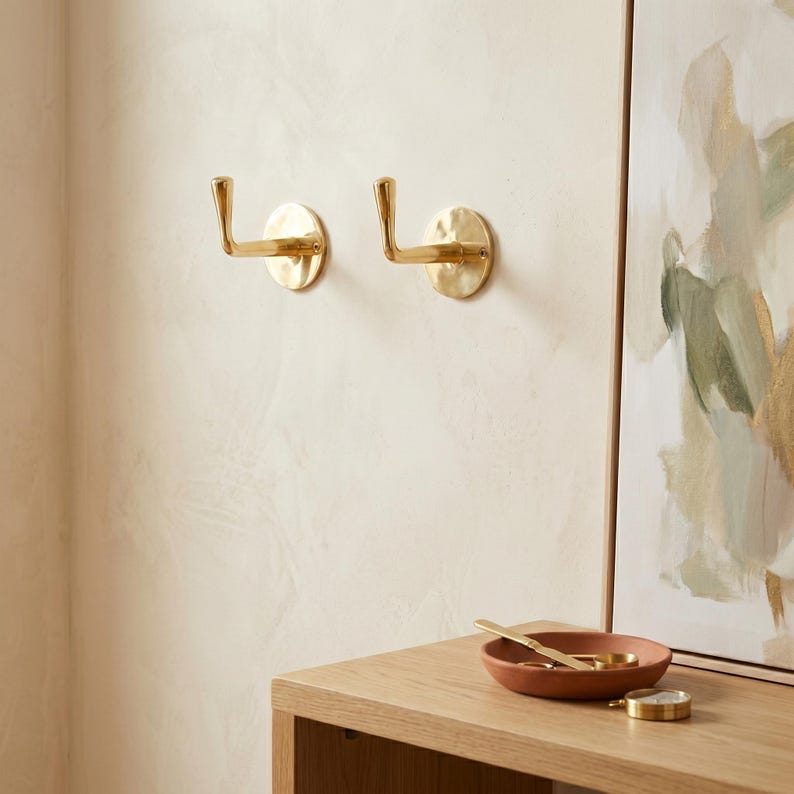 May include: Two brass wall hooks with a hammered finish are mounted on a light-colored wall. The hooks have a simple, elegant design, perfect for hanging coats, bags, or other items. A small, round, terracotta-colored bowl and a gold-colored watch are on a wooden surface.