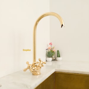 May include: A gold-toned kitchen tap with a curved spout and two cross-shaped handles. The tap is mounted on a white worktop.