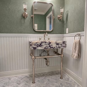 May include: A bathroom vanity featuring a white and purple marble sink basin supported by a polished silver metal frame. The setup includes a silver faucet, a rectangular mirror, two silver wall sconces, and a beige towel on a ring.
