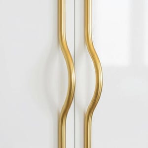 Peut inclure: Deux poignées de placard dorées et ondulées sur fond blanc. Les poignées ont un design lisse et incurvé, ajoutant une touche moderne aux meubles. La quincaillerie est en métal et a une finition polie.