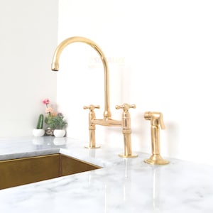 Może przedstawiać: Złota bateria kuchenna z wysoką wylewką w kształcie łuku i krzyżowym uchwytem. Bateria zawiera spryskiwacz boczny. Armatura jest ustawiona na białym marmurowym blacie i białej ścianie. Mała roślina doniczkowa jest widoczna w tle.