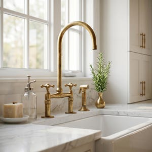 Puede incluir: Un grifo de cocina dorado con un caño de arco alto y un diseño de manija en cruz. El grifo está montado en una encimera blanca, con un pulverizador lateral a juego. También se ven un dispensador de jabón de vidrio transparente y un pequeño jarrón con vegetación.