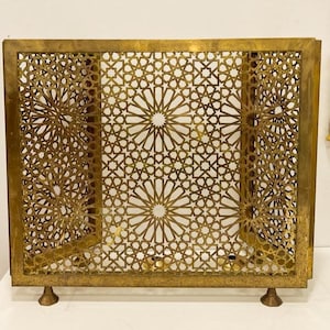 May include: A brass-toned decorative screen featuring a detailed geometric star pattern. The rectangular frame supports the screen, which is adorned with repeating star and floral motifs. The screen stands on four small feet, creating an ornate and visually striking design.