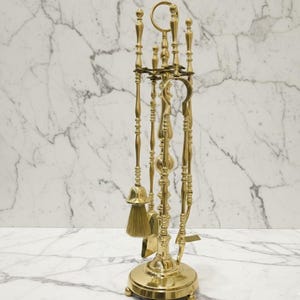 Puede incluir: Un juego de herramientas para chimenea de latón con base circular y múltiples herramientas, que incluyen un cepillo, una pala y un atizador. Las herramientas tienen detalles ornamentados y están dispuestas verticalmente. El juego se muestra sobre un fondo de mármol blanco.