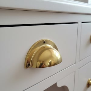 Peut inclure: Gros plan sur un bureau blanc avec une poignée de tiroir de couleur laiton. La poignée de tiroir est en forme de demi-lune et est fixée au tiroir avec deux vis. Le bureau a plusieurs tiroirs et un bord décoratif.