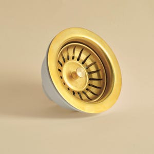 May include: A brass sink strainer with a gold-toned rim and a light grey base. The strainer features vertical slots and a central circular drain. The design is simple and functional, suitable for kitchen or bathroom use.