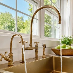 May include: A vintage-style brass kitchen faucet with a high arc spout and cross-handle design. Water flows from the faucet into a gold-toned sink. A side sprayer and a wooden bowl of green apples are also visible.