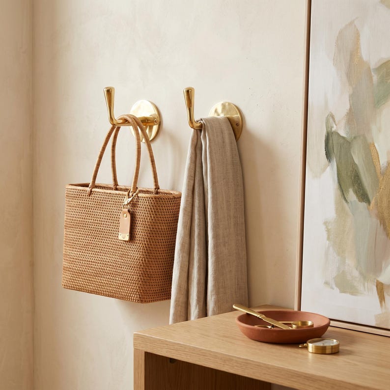 May include: Two brass wall hooks hold a woven tote bag and a light-colored scarf. The bag is rectangular with a leather tag. A small terracotta bowl and a gold-colored compass sit on a wooden surface.