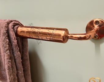 May include: A copper towel bar with an ornate design. The bar is curved and has a round base. A pink towel is hanging on the bar.