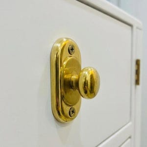 Peut inclure: Gros plan sur une armoire blanche avec un bouton et une plaque arrière dorés. Le bouton est rond et la plaque arrière est ovale, fixée avec deux vis. La porte de l'armoire est blanche avec un panneau encastré.