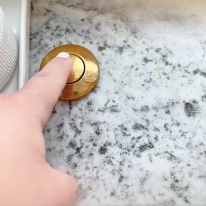 May include: A close-up of a gold-colored, circular button being pressed by a finger. The button is set into a speckled, light-colored surface. The button appears to be part of a fixture.
