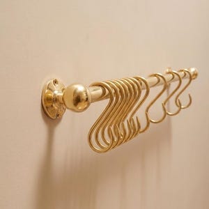 Peut inclure: Un rail de cuisine en métal doré avec une série de crochets en forme de S. Le rail est fixé au mur et comporte des embouts décoratifs. Les crochets sont espacés uniformément, offrant une solution de rangement élégante pour les ustensiles de cuisine. La longueur du rail est d'environ 30 cm.