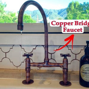 Puede incluir: Un grifo de puente de cobre con manijas en cruz. El grifo tiene un caño curvo y está montado en una encimera. El texto "Copper Bridge Faucet" se muestra encima del grifo. A la derecha hay una botella de vidrio oscuro de jabón de manos.