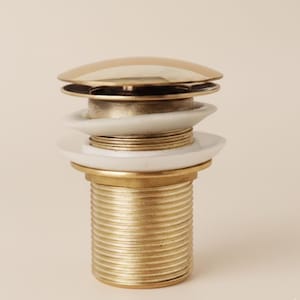 May include: A close-up of a gold-colored pop-up drain stopper. The stopper has a shiny, rounded top and a threaded base. White ceramic rings and brass components are visible.