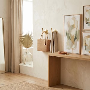 May include: A woven tote bag with a tag hangs on a gold hook on a cream-colored wall. A beige towel hangs on a second gold hook. A wooden table holds a small bowl and artwork.