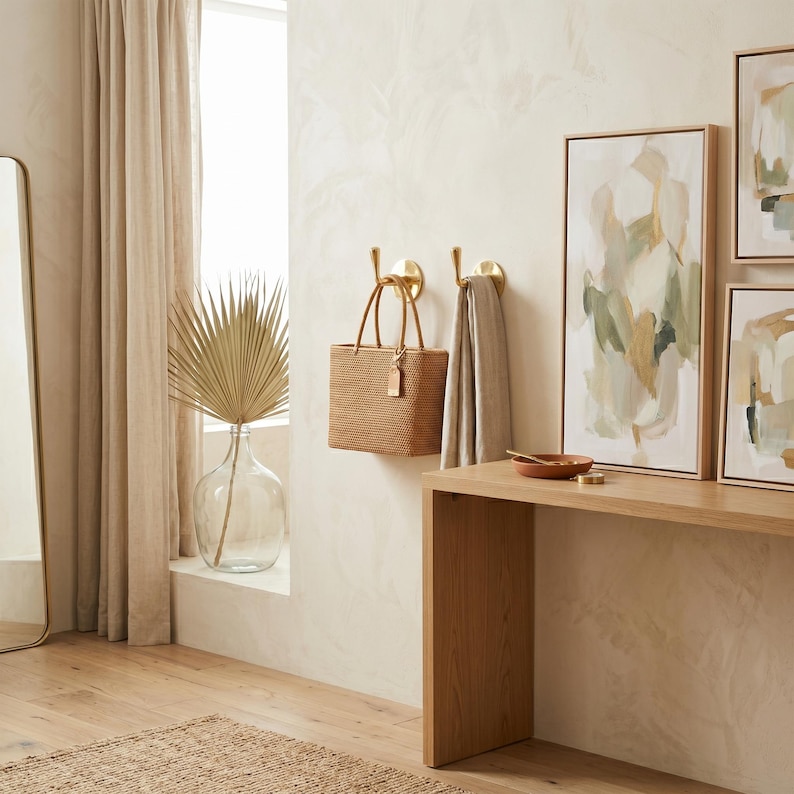 May include: A woven tote bag with a tag hangs on a gold hook on a cream-colored wall. A beige towel hangs on a second gold hook. A wooden table holds a small bowl and artwork.