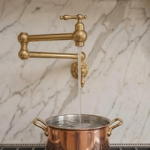 Op de afbeelding: Een aan de muur gemonteerde koperen kraan met een dubbel scharnierende zwenkarm en een enkele hendel, die een gestage stroom water in een gepolijste koperen pan met twee handvatten giet, tegen een achterwand van wit marmer.