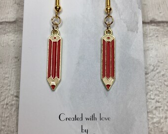 May include: A pair of gold dangle earrings with red and white pencil charms. The earrings are on a white card with the text "Created with love by Absolutely Charming".