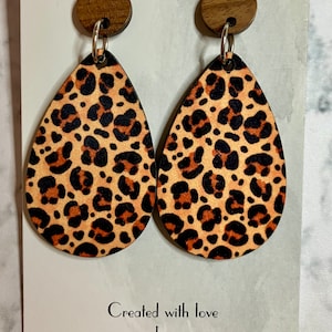 May include: A pair of dangle earrings with a leopard print design. The earrings are made of wood and have a brown wooden top. The earrings are on a white background with the text "Created with love by Absolutely Charming".