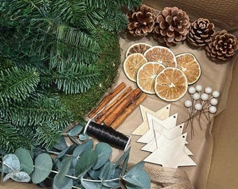 May include: A box of Christmas wreath-making materials. It contains a green wreath base, pine cones, dried orange slices, cinnamon sticks, wooden star shapes, eucalyptus, twine, and wire. The items are on brown paper.