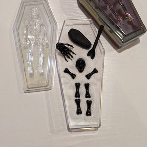 Op de afbeelding: Twee doorzichtige plastic kistvormige containers. Een container heeft een zwart skelet erin. De andere container heeft wit zand, een zwarte schedel, een zwarte hand, een zwarte kist en zwarte botten.