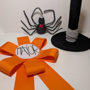 May include: An orange ribbon rosette with a white center circle featuring the word MAYOR in black text, a black spider with silver glitter pipe cleaner legs and a red hourglass mark, and a black felt top hat with a light gray band.