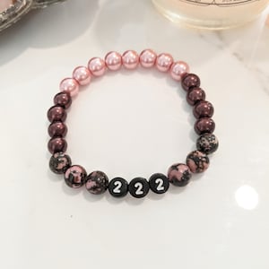以下が含まれることがあります： ピンクとブラウンのビーズブレスレット。黒いビーズで「222」と綴られています。