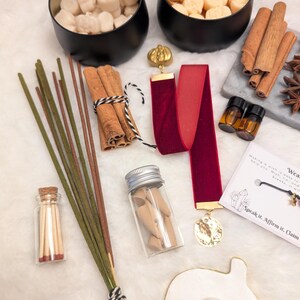 May include: An assortment of aromatherapy and self-care items. Includes black metal bowls with wax melts, cinnamon sticks, incense, essential oil bottles, a red velvet ribbon with a gold pendant, and a small glass jar with wooden figurines.