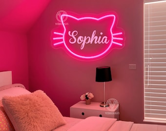 Könnte beinhalten: Rosafarbenes Neon-Schild in Form eines Katzenkopfes mit dem Namen "Sophia" darin. Das Schild ist an einer rosa Wand in einem Schlafzimmer mit einem weißen Bett und einer schwarzen Lampe auf einem weißen Nachttisch angebracht.