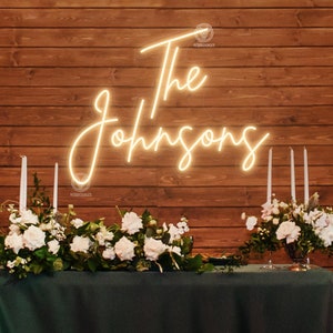 May include: A white neon sign with the text "The Johnsons" on a wooden wall background. The sign is illuminated and is placed above a table with white flowers and candles.