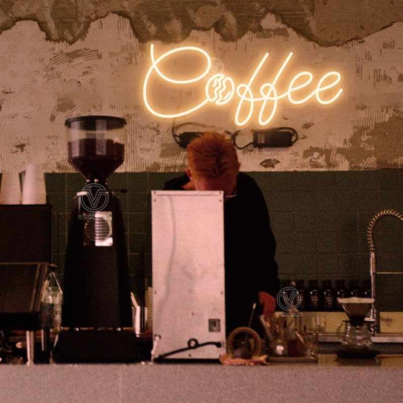 May include: A neon sign with the word "Coffee" in a cursive font, with a stylized coffee cup and steam design. The sign is lit up in a warm yellow colour and is hanging on a wall with a distressed, textured surface.