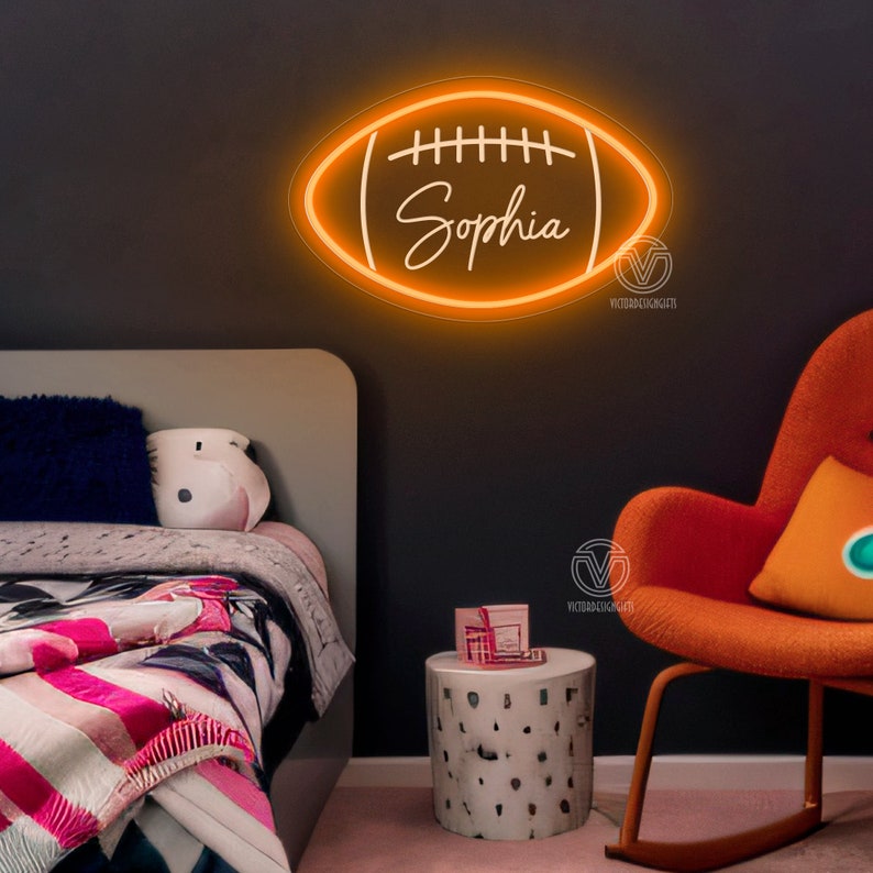 May include: Orange neon sign shaped like a football with the name "Sophia" inside. The sign is hanging on a dark grey wall in a bedroom with a bed, a chair, and a side table.