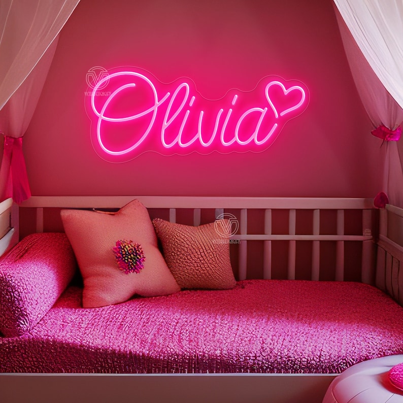 K&ouml;nnte beinhalten: Ein leuchtend pinkes Neonschild an einer Wand, das den Namen Olivia in Schreibschrift zeigt, begleitet von einem Herzsymbol. Das Schild befindet sich &uuml;ber einem Bett mit pinker Bettw&auml;sche, einem strukturierten pinken Kissen und einem kleineren Kissen.